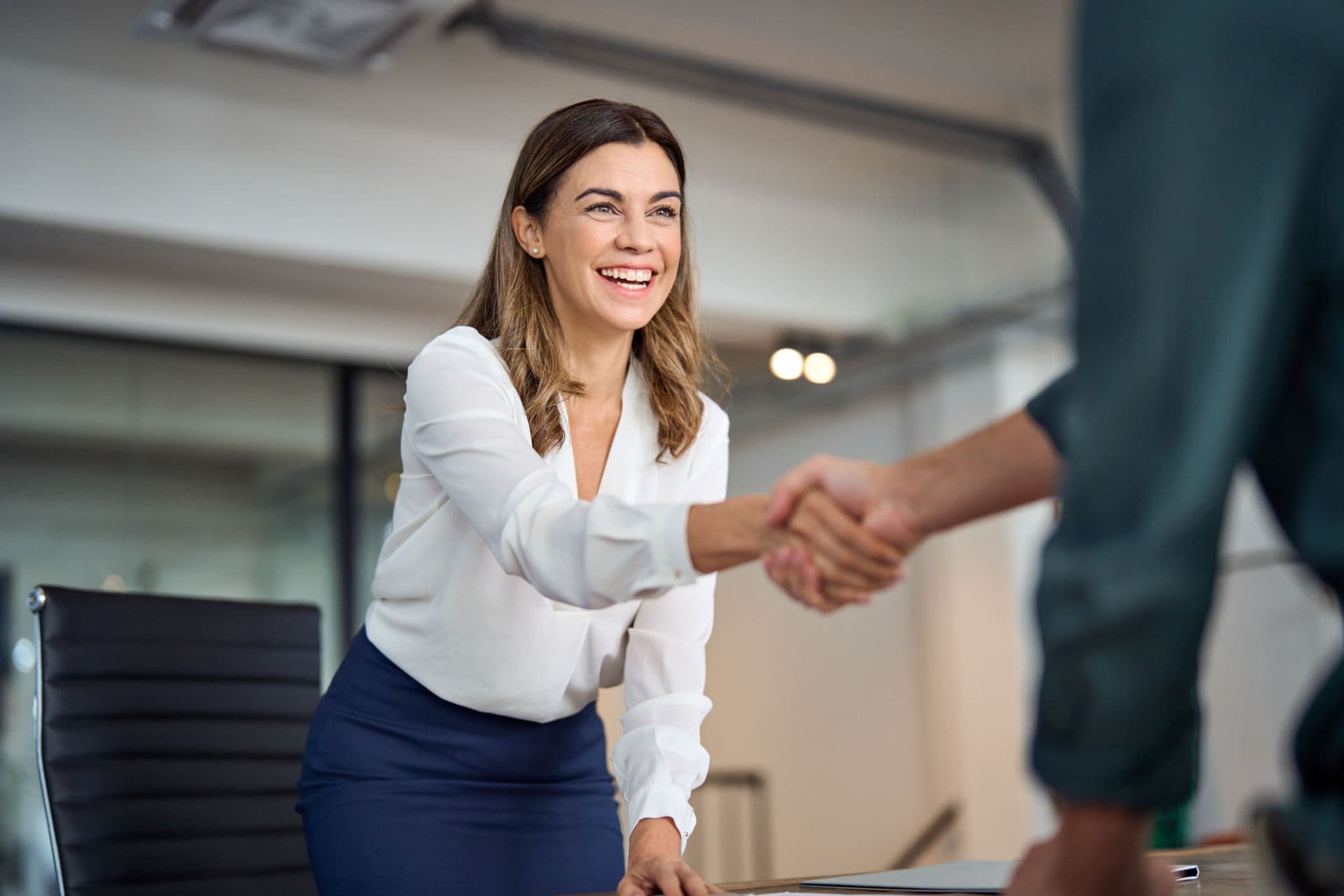 Smiling businesswoman in a white blouse shaking hands with a colleague in a modern office.