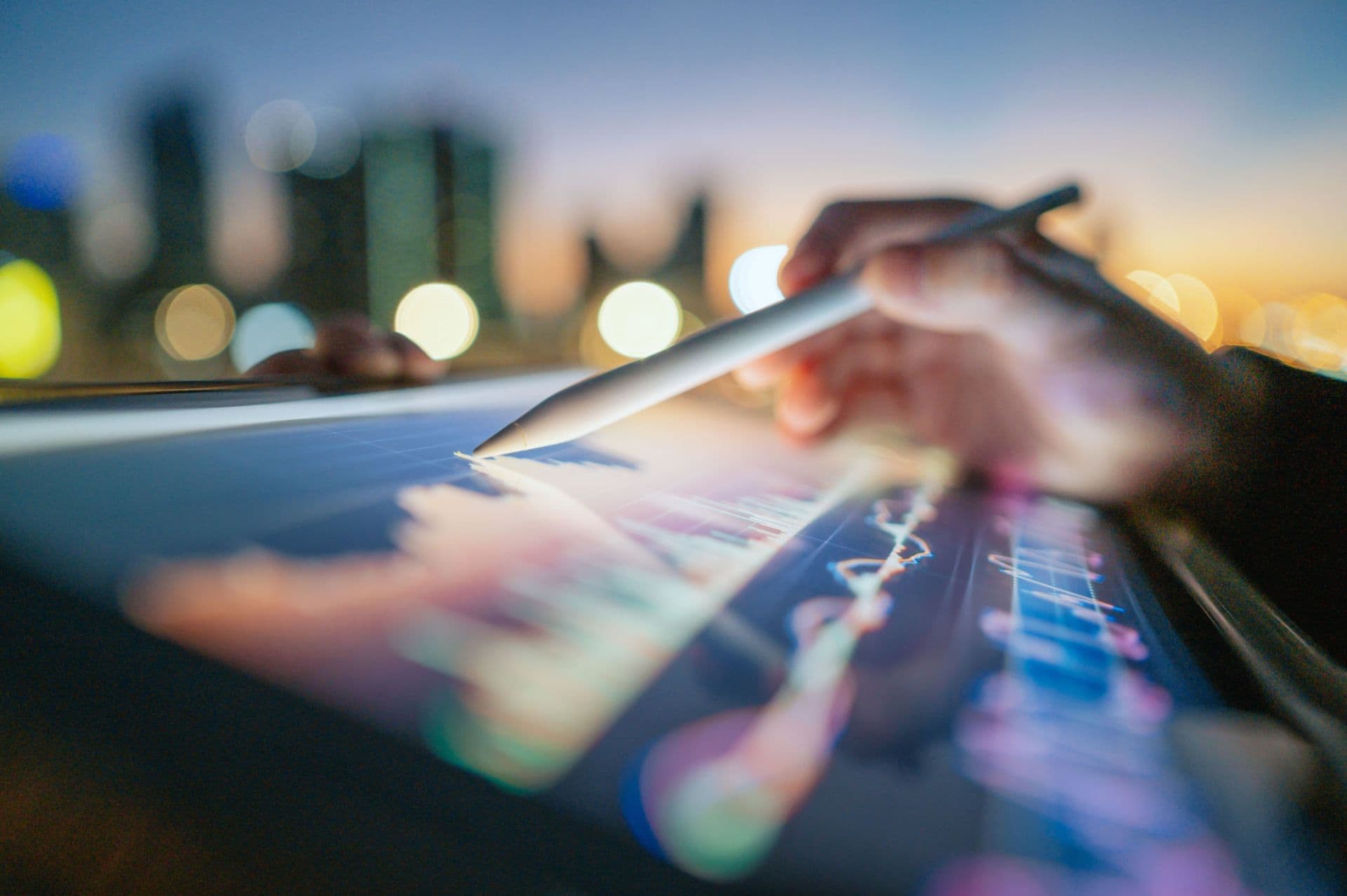 Hand using a stylus to analyze financial charts on a tablet against blurred city lights.